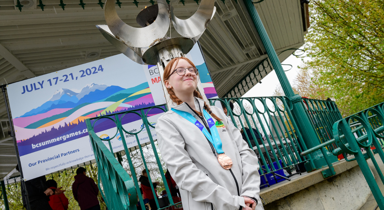 A young athlete poses with her medal in front of a big BC Games torch.